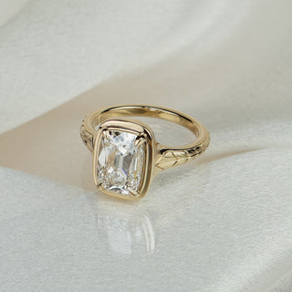 Old mine cushion cut lab grown diamond engagement ring in yellow gold on satin fabric, bezel set center over a hand-engraved shank.