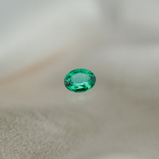 A single oval-cut emerald green gemstone with faceted edges, resting on a soft, light beige textured surface.