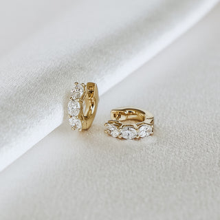 A pair of small gold huggie hoop earrings with clear oval gemstones, displayed on a soft white satin fabric background.