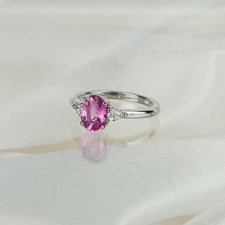 Oval pink Sapphire three-stone engagement ring in white gold on satin fabric, pear shape Diamonds flanking the center.