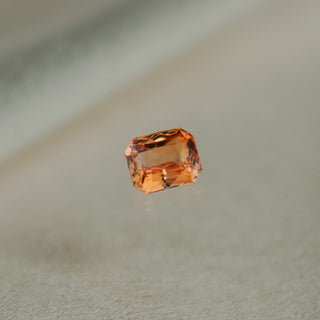 A single rectangular emerald-cut orange gemstone with faceted edges, centered on a soft, light beige textured surface.