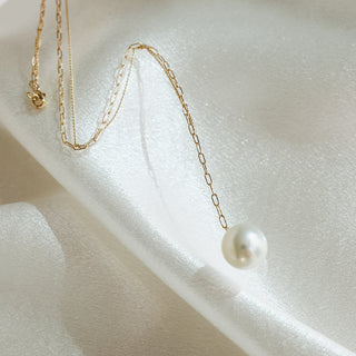 A delicate gold chain necklace with a single white pearl pendant, elegantly draped on a soft, textured white satin fabric.