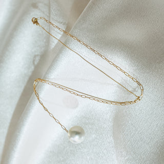 A delicate gold chain necklace with a single white pearl pendant, laid out on a soft, white satin fabric background.