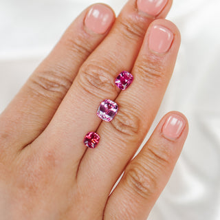 Three pink gemstones of varying sizes arranged vertically on a person's index finger against a soft white background.