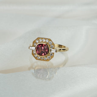 Cushion cut pink Spinel engagement ring in yellow gold on satin fabric, geometric milgrain frame with Diamonds and tapered baguettes.