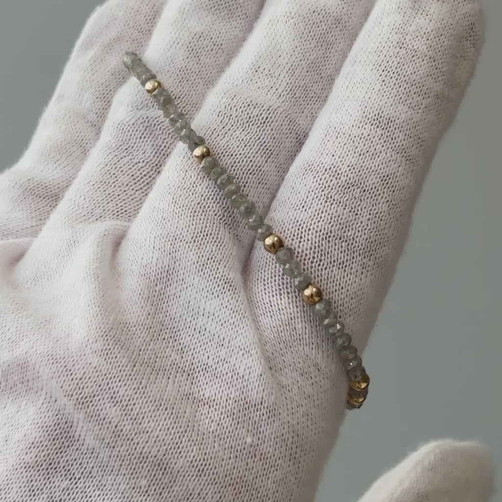 Video of a natural grey raw faceted Diamond bracelet draped over a gloved hand, grey stones and gold beads in motion.