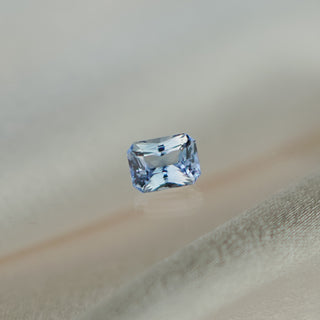 A single rectangular cushion-cut light blue gemstone with faceted edges, resting on a soft beige satin fabric surface.