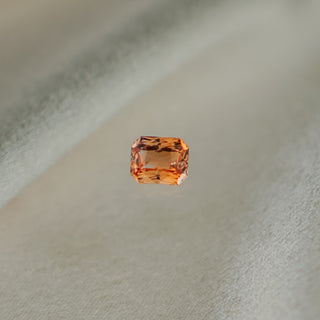 A single rectangular emerald-cut orange gemstone with faceted edges, centered against a soft, light beige textured fabric background.