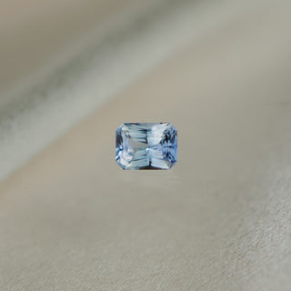 A single rectangular emerald-cut light blue gemstone with faceted clarity, centered on a soft beige textured background.