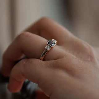 Natural teal Sapphire engagement ring worn on the hand, emerald cut center shifting between green and blue.