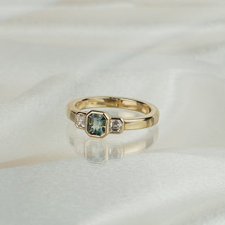 Emerald cut teal Sapphire three-stone engagement ring in yellow gold on satin fabric, bezel set center with Asscher cut Diamond sides.