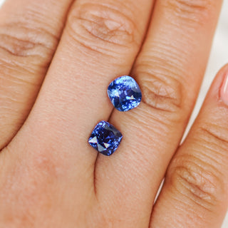 Two loose cushion-cut blue gemstones displayed on a person's fingers against a light neutral background.