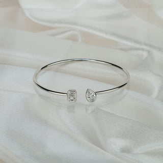 Lab grown diamond toi et moi bangle in white gold on satin fabric, emerald cut and pear shape stones facing each other at the open ends.