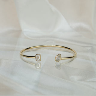 Open yellow gold bangle with emerald cut and pear lab grown diamonds lying on ivory satin, cuff gap facing forward.