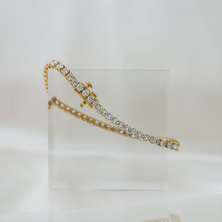 Yellow gold tennis bracelet with round brilliant natural Diamonds, strand curving over a clear block against soft fabric.
