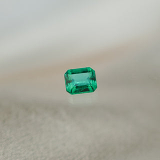 A single rectangular emerald-cut emerald, centered on a soft, light beige textured surface with a blurred neutral background.