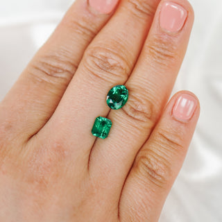 A close-up of a hand wearing a pair of green gemstone stud earrings, with one oval-cut green stone and one rectangular-cut green stone on a silver-toned setting.